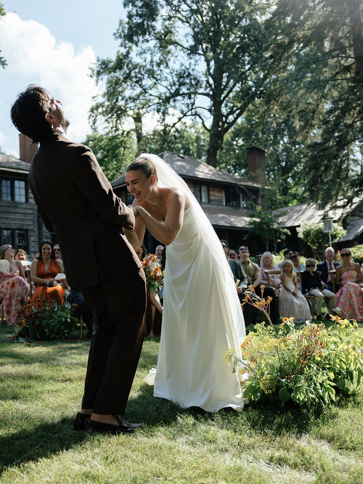 A groom throwing his head back laughing at the altar while his bride doubles over beside him, guests laughing in the rows behind