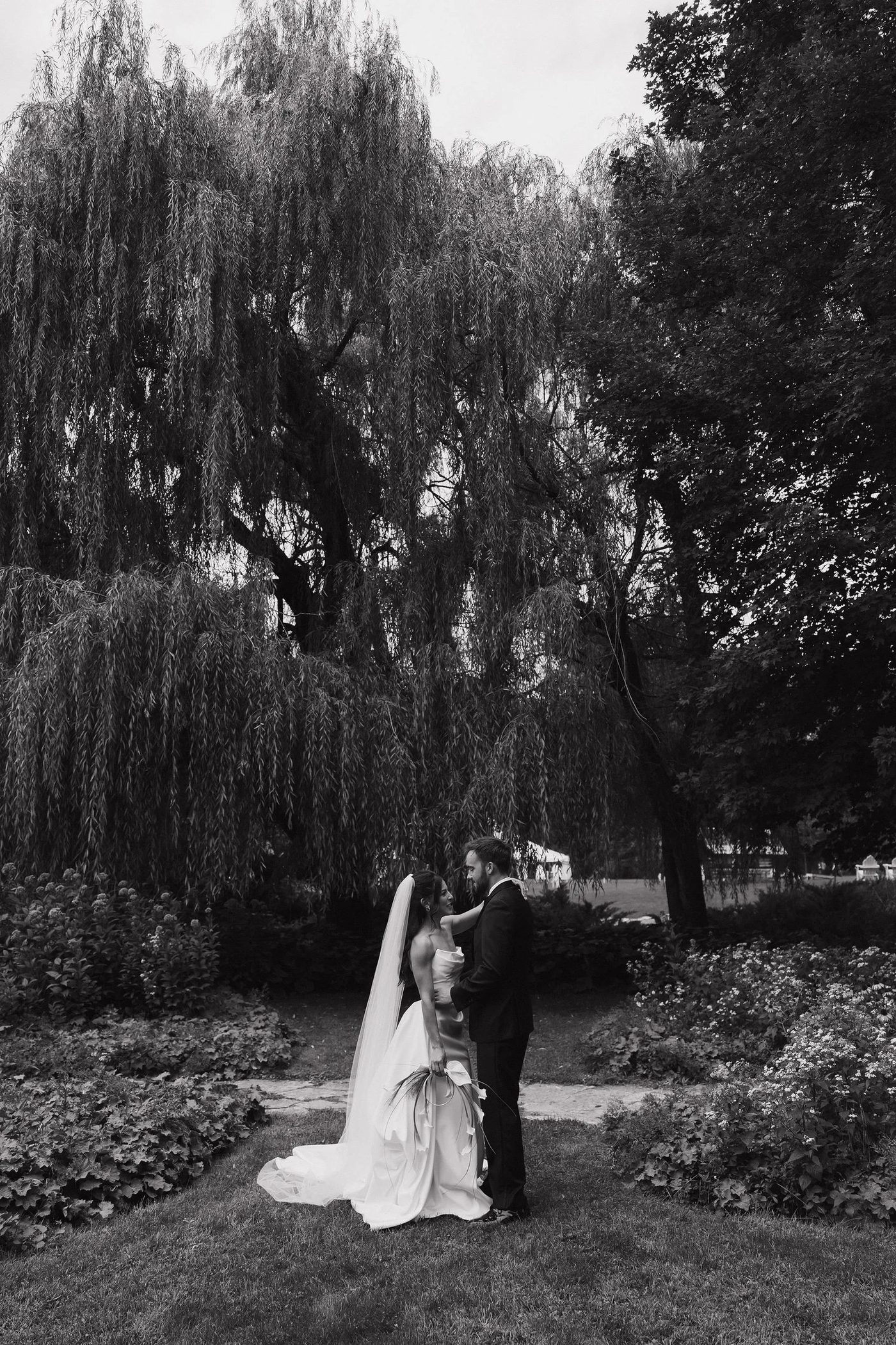 Maddy and Matt beneath a sweeping willow tree, her veil trailing through the garden — black and white