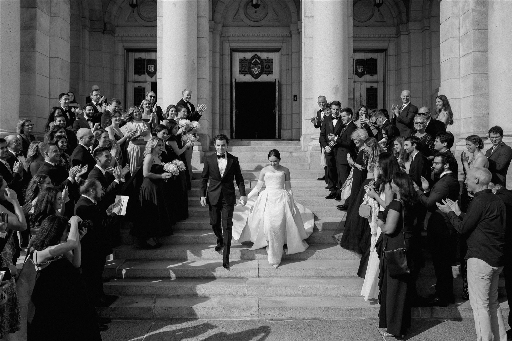 Rachel and Peter descending the Basilica steps hand in hand through two rows of applauding guests, in black and white