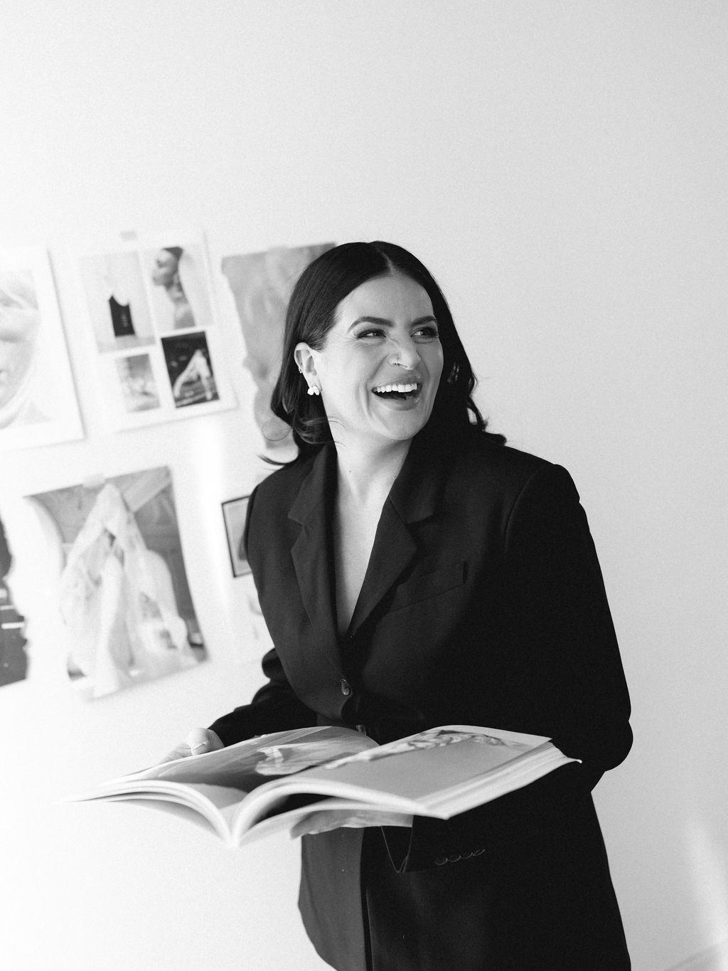 Steph laughing in the studio, an open magazine in her hands and her moodboard wall behind her, in black and white