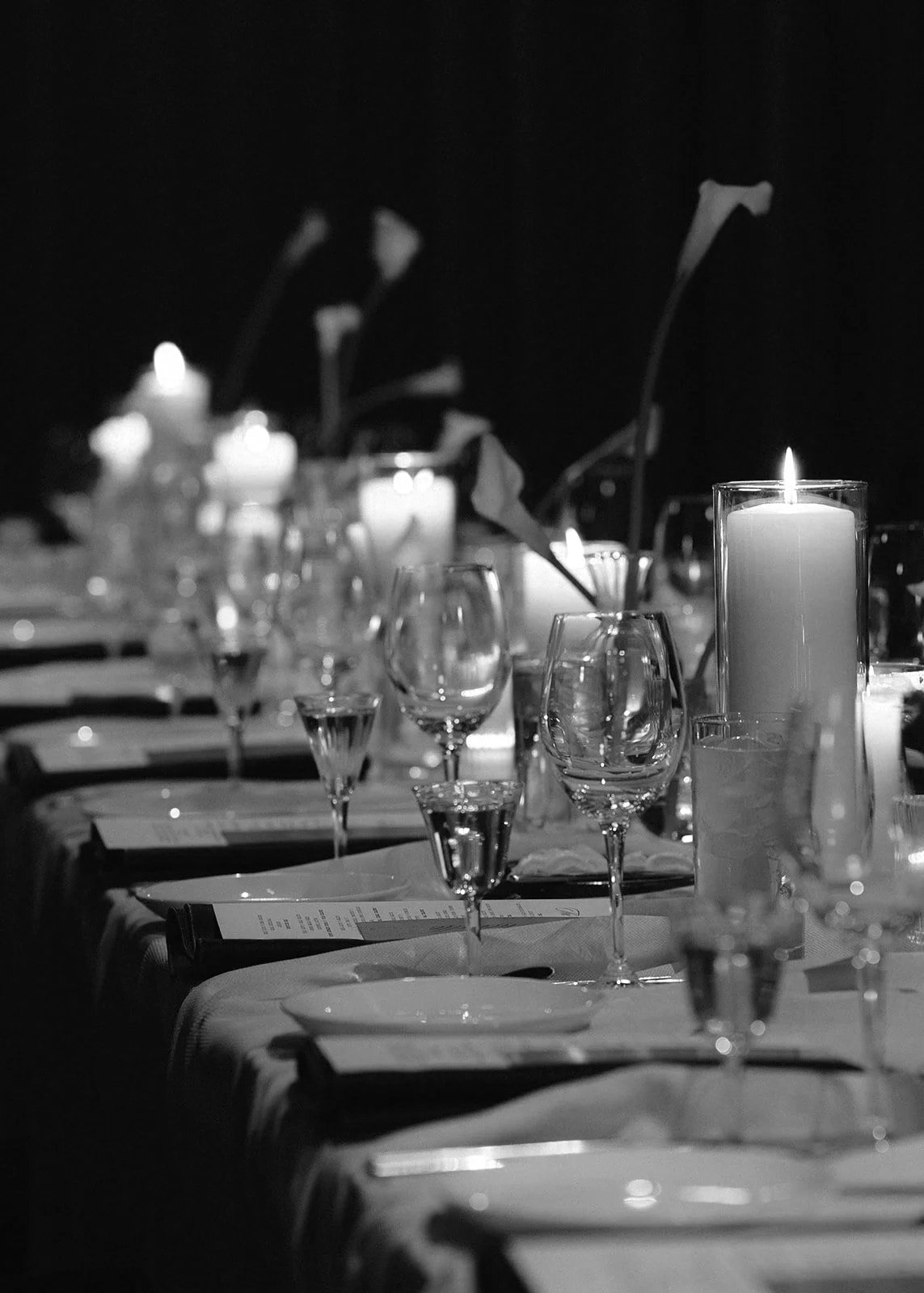 Candlelight and glassware down a black-tie dinner table, in black and white