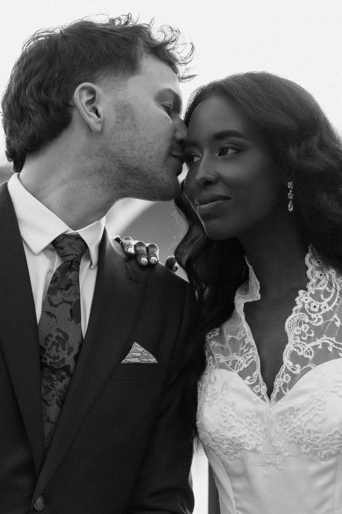 A couple close together, his kiss against her temple, her hand resting on his lapel, in black and white