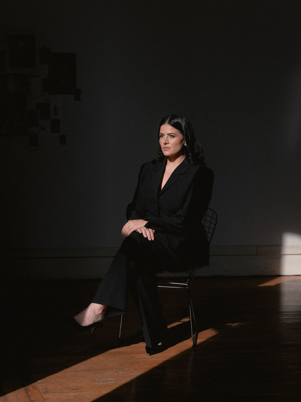 Steph seated in a shaft of warm light in a darkened studio, her moodboard wall behind her